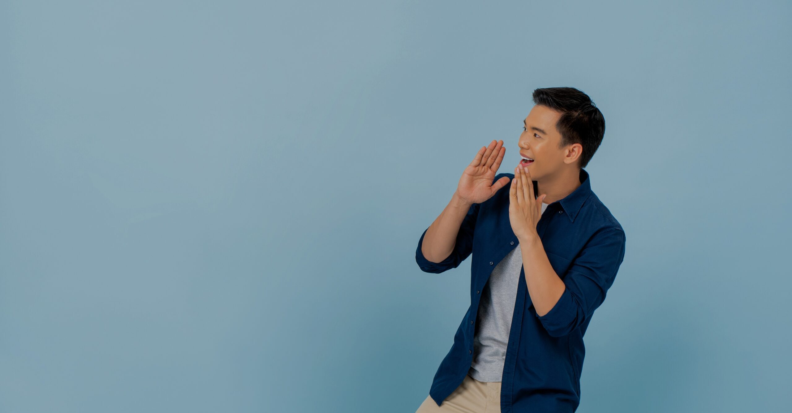 Young Asian man shouting and cupping hands around mouth in light blue isolated background with copy space