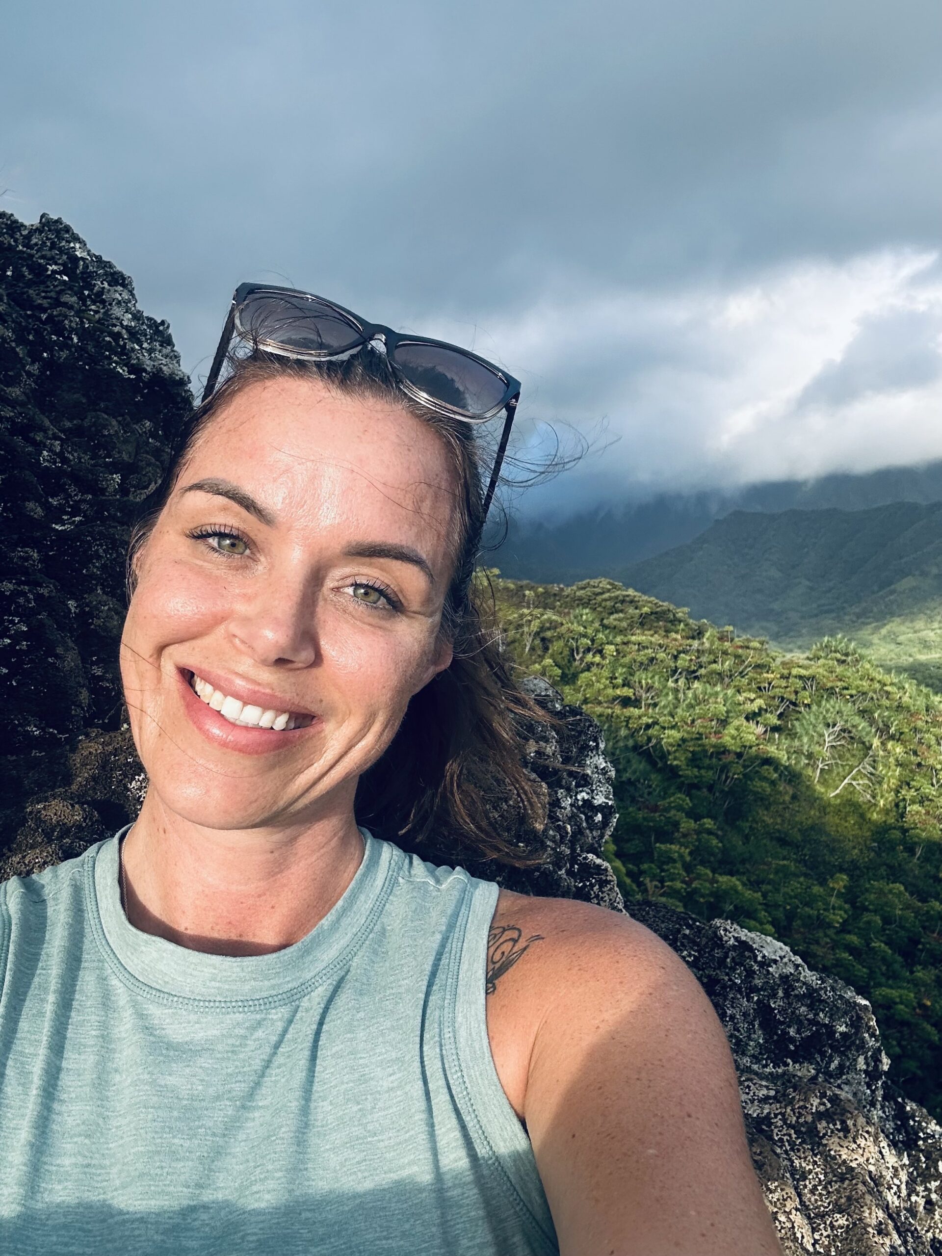 Samantha Behiry smiles for a selfie in a light blue sleeveless top, perched on a rocky outcrop with lush green mountains and a cloudy sky behind her. Sunglasses rest casually on her head.