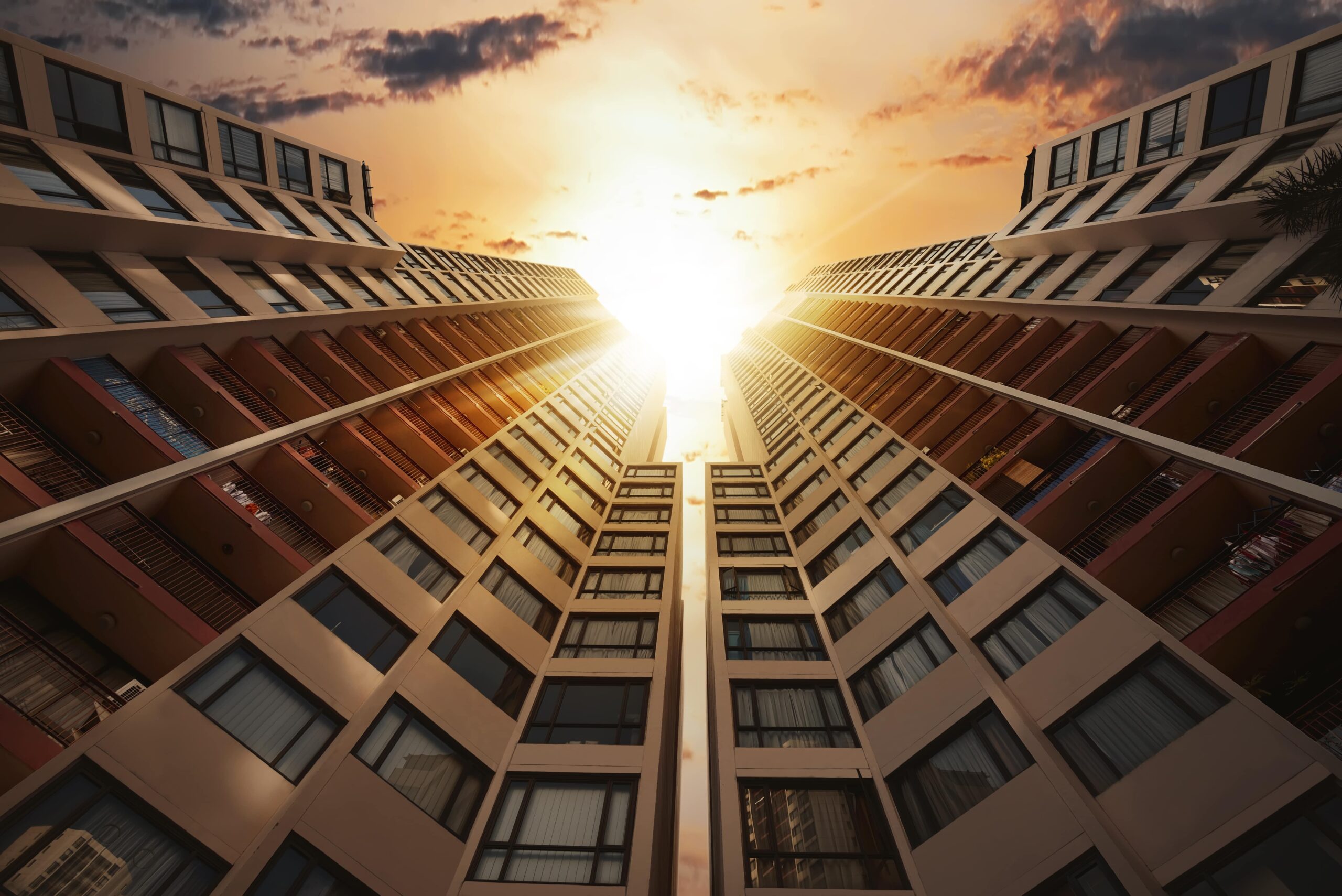 Two tall apartment buildings rise on either side, converging toward the sky, with a bright sun setting between them and casting a golden glow. The perspective is from ground level, looking upward.