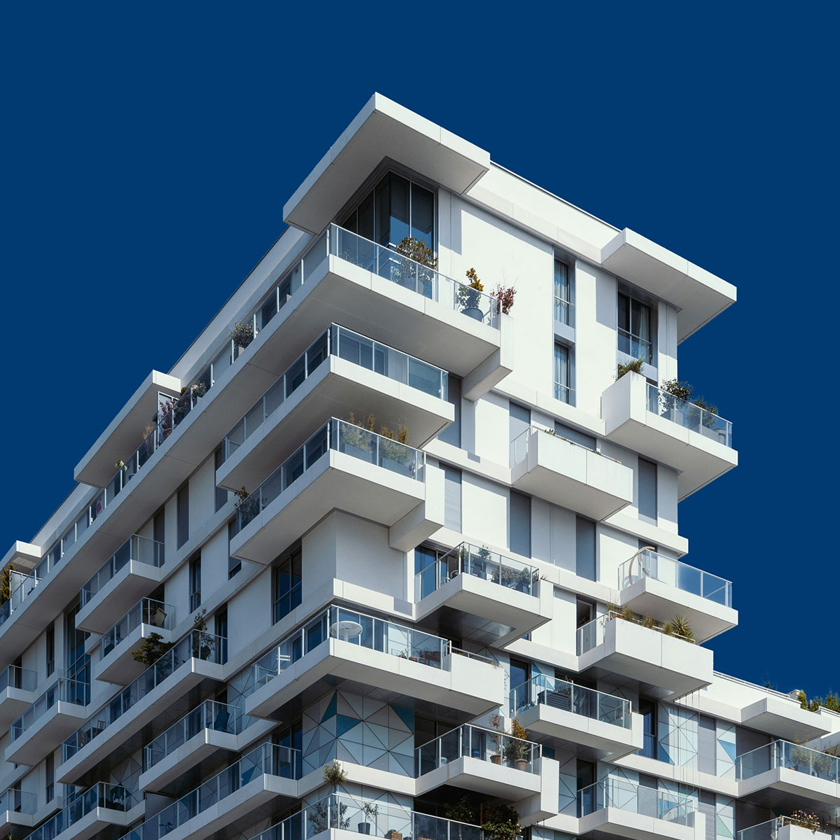 A modern white apartment building with multiple balconies and glass railings, featuring plants and outdoor furniture, set against a clear, deep blue sky.