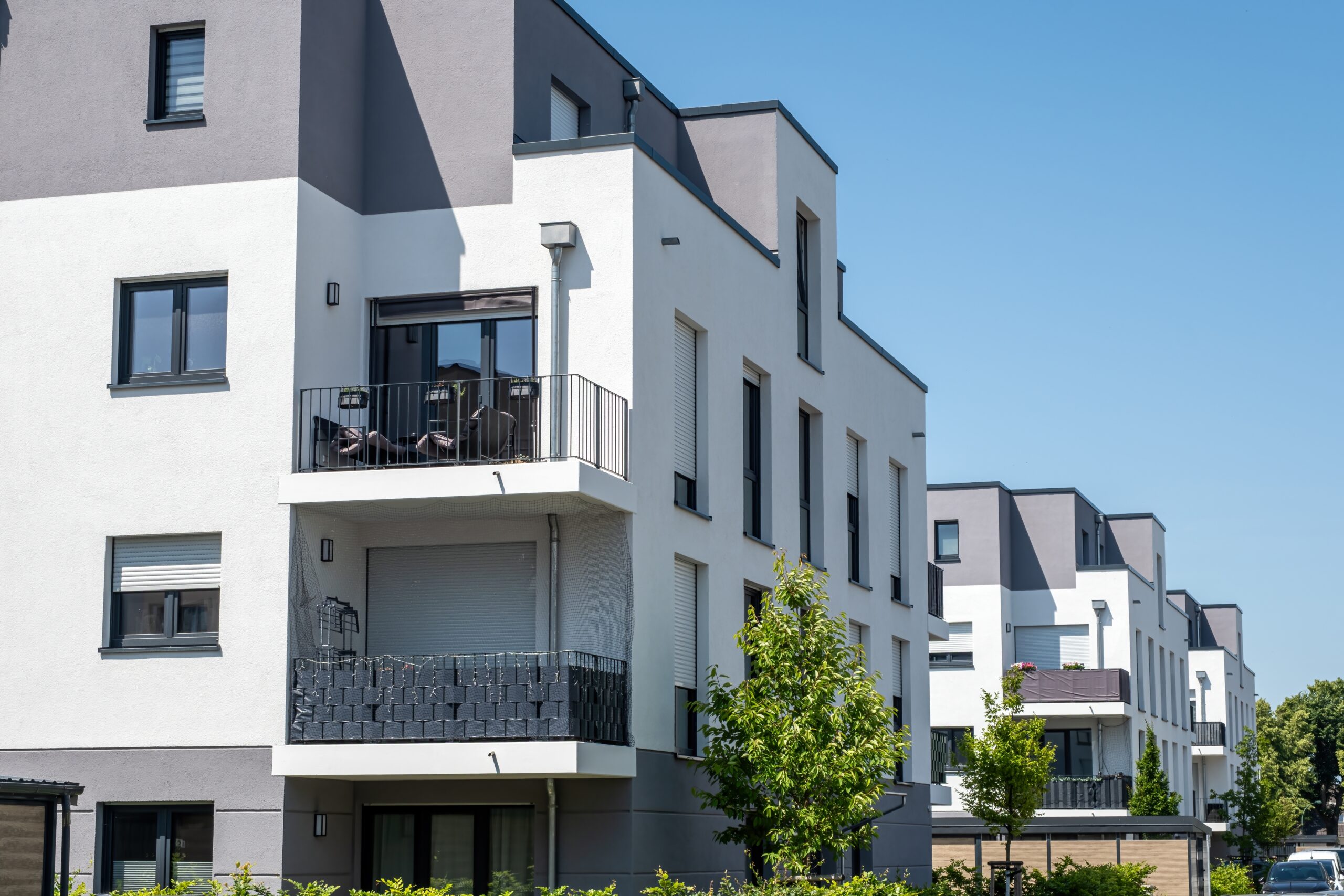 Modern white and gray apartment buildings with balconies and large windows rise against a clear blue sky, surrounded by green trees—designed to boost multifamily occupancy in today’s competitive markets.