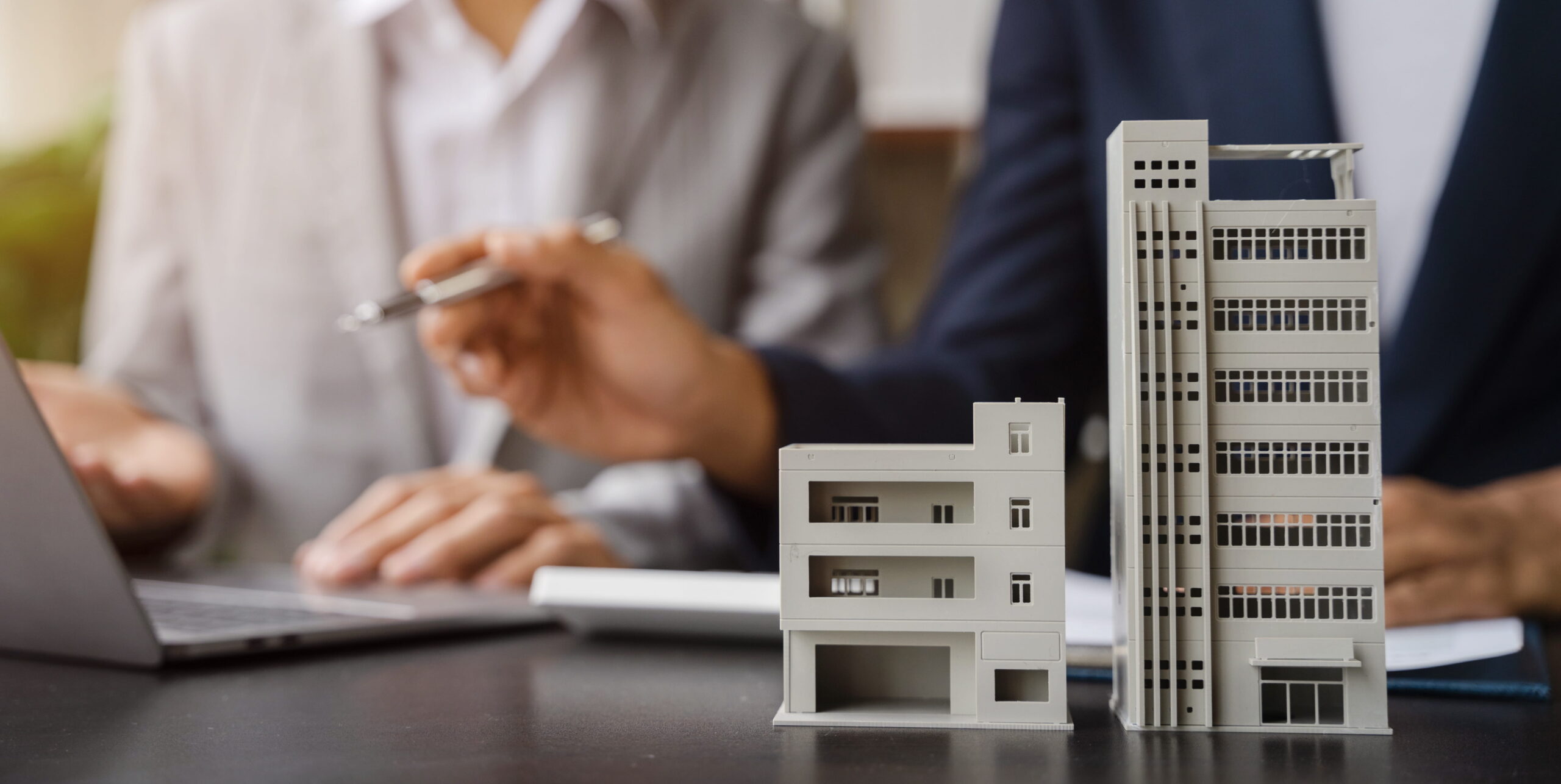 Two people in business attire sit at a desk with a laptop, discussing financial analysis. In the foreground are two model buildings, representing real estate or property investment and the calculation of net operating income (NOI).